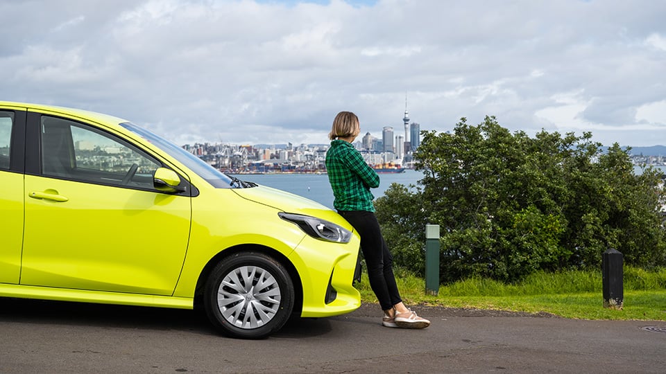 A person leaning on the front of a Toyota Yaris looking across the water at Auckland CBD